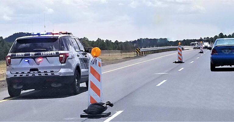 A state trooper vehicle with its lights on, parked on a highway shoulder behind traffic cones